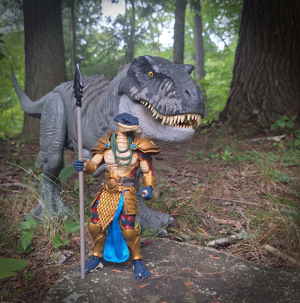 Toy photo of Varnakk, a Snake-Man warrior, standing with spear in hand beside his companion T-Rex. The dinosaur looms behind him, wide-eyed as if catching a scent, while Varnakk waits in silence, poised to strike. The scene captures the anticipation of a mythic hunt in a forest setting.